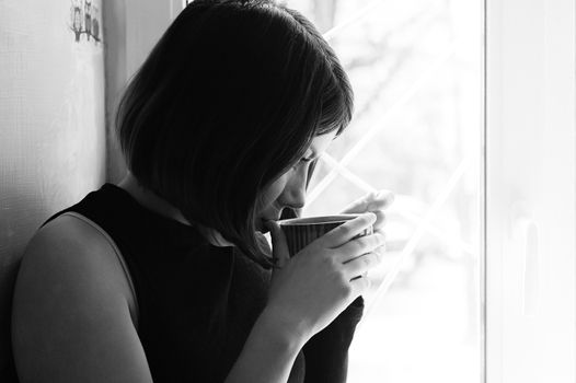black and white portrait of a young brunette girl in black with a big cup of tea by the window