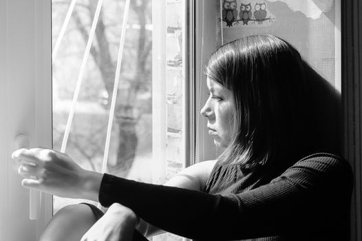 black and white image of a sad young pretty brunette woman in black clothes looking out the window