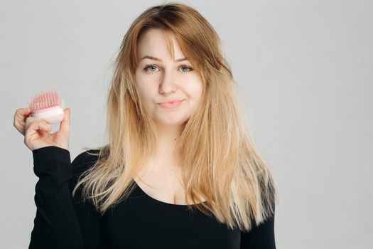 Close up portrait of young confused girl with messy blonde hair. Pretty woman with green eyes, wearing in black sweater, holding hairbrush in hand and looking at camera against white background.