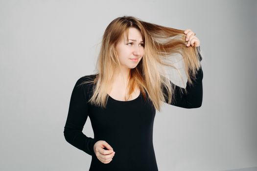 Pretty blonde haired woman wearing in black sweater, holding messy unbrushed hair in one hand and pink hairbrush in another one. A frustrated girl looks at her hair on a white studio background.