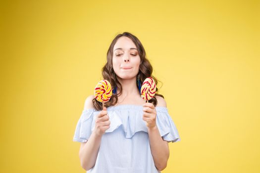 Close up portrait of attractive lovely girl in light dress handing lolipop isolated on yellow background