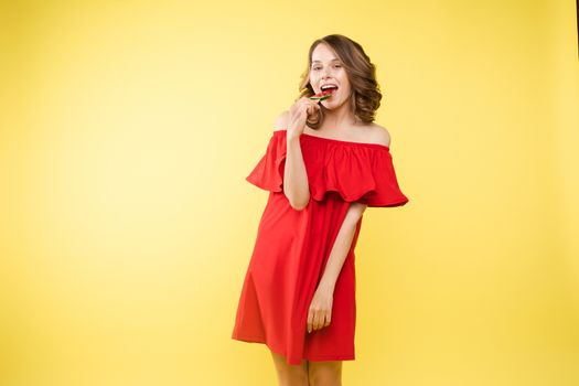 Beautiful and seductive woman wearing like sweet doll, posing at studio with candy on stick. Confident pretty woman in dress andsunglasses, holding hand on waist. Fashion, glamour.