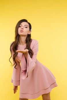 Beautiful young girl wearing short pink dress and trendy hairstyle looking at camera and posing on yellow isolated background. Attractive woman modeling in studio. Concept of fashion and beauty.