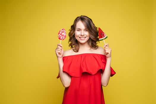 Close up portrait of attractive lovely girl in light dress handing lolipop isolated on yellow background