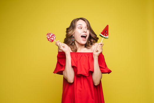 Beautiful and seductive woman wearing like sweet doll, posing at studio with candy on stick. Confident pretty woman in dress andsunglasses, holding hand on waist. Fashion, glamour.