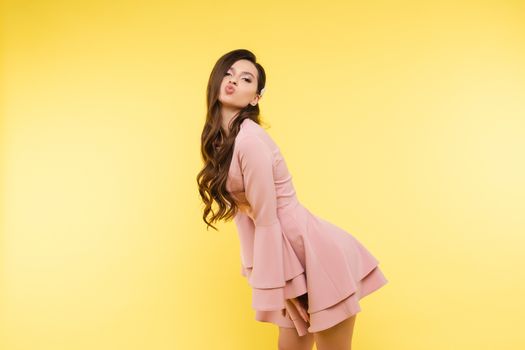 Side view of brunette girl with long hair posing at camera. Young beautiful girl in pink dress lightly leaning forward and flirting. Pretty glamour lady sending in elegant and stylish look air kisses.