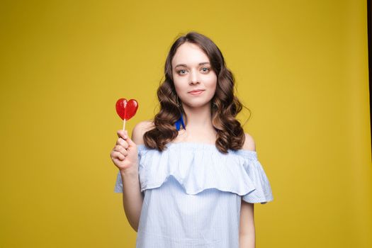 Beautiful and seductive woman wearing like sweet doll, posing at studio with candy on stick. Confident pretty woman in dress andsunglasses, holding hand on waist. Fashion, glamour.