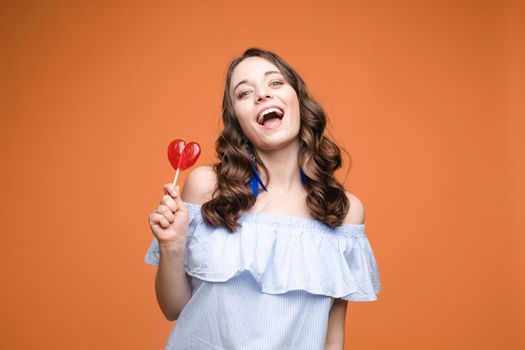 Beautiful and seductive woman wearing like sweet doll, posing at studio with candy on stick. Confident pretty woman in dress andsunglasses, holding hand on waist. Fashion, glamour.