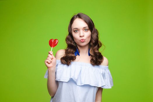 Beautiful and seductive woman wearing like sweet doll, posing at studio with candy on stick. Confident pretty woman in dress andsunglasses, holding hand on waist. Fashion, glamour.