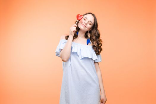 Beautiful and seductive woman wearing like sweet doll, posing at studio with candy on stick. Confident pretty woman in dress andsunglasses, holding hand on waist. Fashion, glamour.