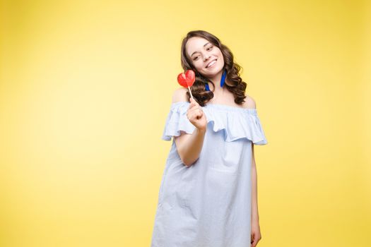 Close up portrait of attractive lovely girl in light dress handing lolipop isolated on yellow background