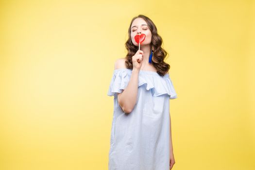 Beautiful and seductive woman wearing like sweet doll, posing at studio with candy on stick. Confident pretty woman in dress andsunglasses, holding hand on waist. Fashion, glamour.