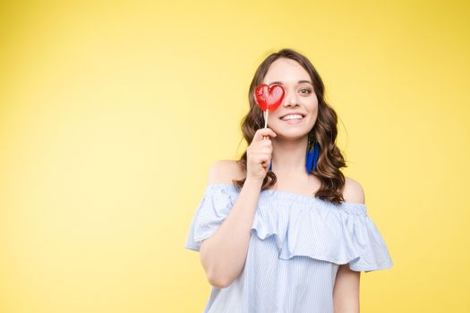 Beautiful and seductive woman wearing like sweet doll, posing at studio with candy on stick. Confident pretty woman in dress andsunglasses, holding hand on waist. Fashion, glamour.