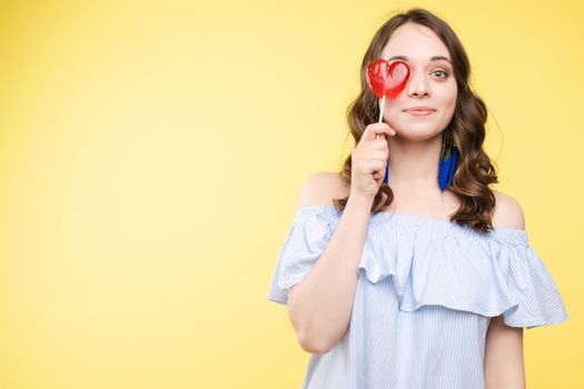 Close up portrait of attractive lovely girl in light dress handing lolipop isolated on yellow background