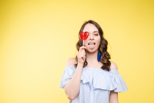 Beautiful and seductive woman wearing like sweet doll, posing at studio with candy on stick. Confident pretty woman in dress andsunglasses, holding hand on waist. Fashion, glamour.