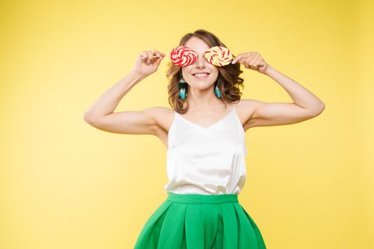 Full length studio portrait of laughing model in white top and green skirt and heels holding two sweet candies on her eyes like sunglasses with her mouth open. Isolate on yellow.