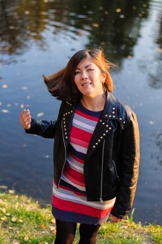 cute young woman with red hair in a leather black jacket and striped color sweater smiles near the water