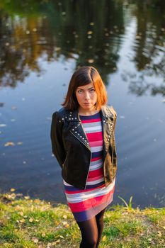 nice European black-haired girl in a multi-colored knitted dress and a black leather jacket stands near the water