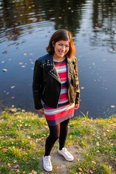 pretty fashionable young woman in a colorful striped sweater and black leather jacket stands on the shore