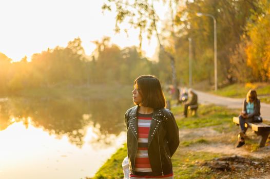 young brunette girl in a black jacket and striped t-shirt looks at the lake in the park