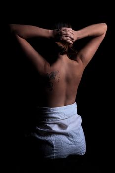 young girl with dark hair and a tattoo stands with naked back in the dark, holding her hands on her head