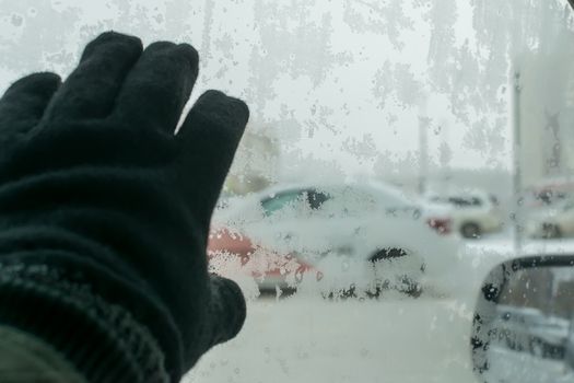 the hand of a man in a winter glove touches the icy glass of the car, on which snow patterns, snowflakes, on the background of the cityscape. Cold in the car