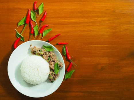 A white plate with steamed rice and stir fried pork with basil Beside, there are chilli and freckles because they are beautifully decorated. The top view on the background is hardwood.