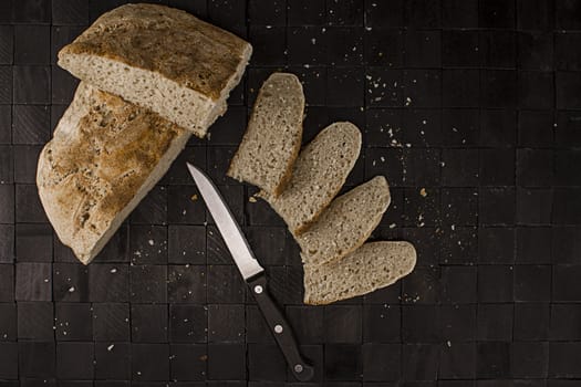 Homemade bread, simple and healthy nutrition. The loaf of bread cut with various slices and a knife, seen from above on a black background