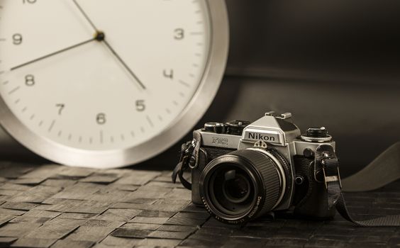 The frenzy of today's times in contrast with the past. A modern design wall clock next to a vintage camera on a black background with a sepia effect