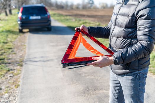 young man installs an orange warning triangle on the road in front of a broken car