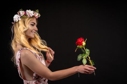 portrait of a young woman in a dress on a black background with a floral wreath on her head. she brought a red rose in her hand