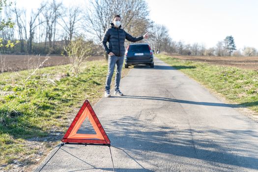 red warning triangle on the road in front of a broken car. The young man is looking for help stopping vehicles moving around.