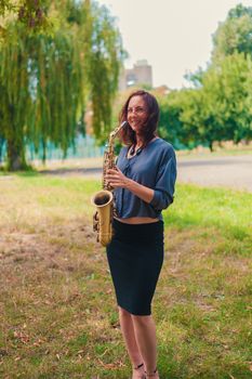 cute young redhead woman in blue blouse and black skirt posing with saxophone in the park