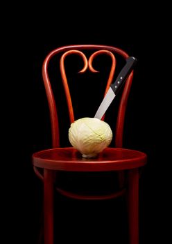 Symbolic conceptual image of a white cabbage on a wooden Thonet chair. A knife is stuck into the vegetable and symbolize cooking, violence, crime, suffering.  On black background.