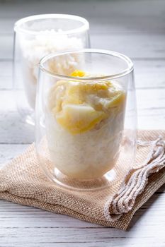 Summer appetizer of Thailand concept. Durian and sticky rice with coconut milk cream in transparent glass on white wooden table and brown table cloth. Famous street food in Thailand.  