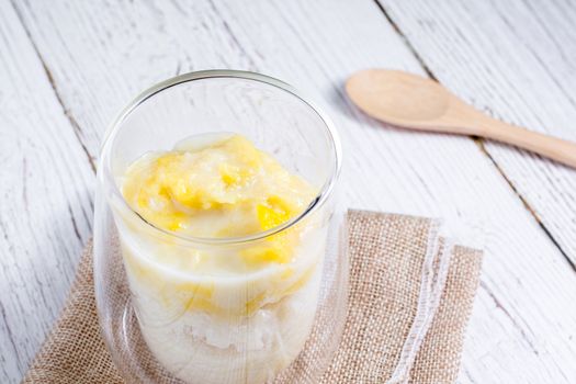 Summer appetizer of Thailand concept. Durian and sticky rice with coconut milk cream in transparent glass on white wooden table and brown table cloth. Famous street food in Thailand.  