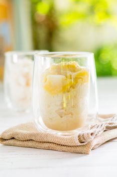 Summer appetizer of Thailand concept. Durian and sticky rice with coconut milk cream in transparent glass on white wooden table and brown table cloth and yellow and green bokeh light background. Famous street food in Thailand.  