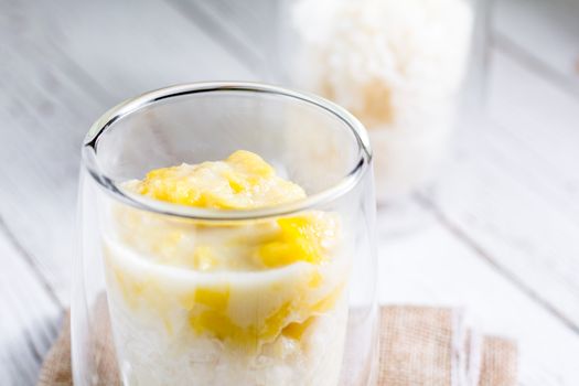 Summer appetizer of Thailand concept. Durian and sticky rice with coconut milk cream in transparent glass on white wooden table and brown table cloth. Famous street food in Thailand.  