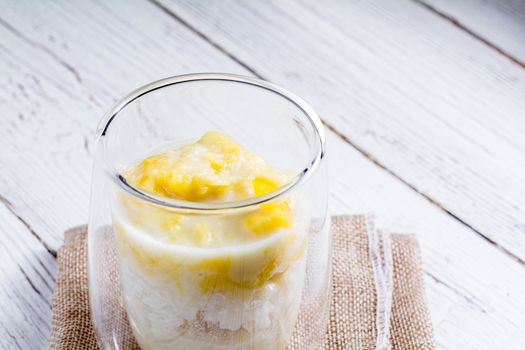 Summer appetizer of Thailand concept. Durian and sticky rice with coconut milk cream in transparent glass on white wooden table and brown table cloth. Famous street food in Thailand.  