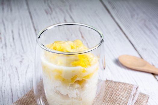 Summer appetizer of Thailand concept. Durian and sticky rice with coconut milk cream in transparent glass on white wooden table and brown table cloth. Famous street food in Thailand.  