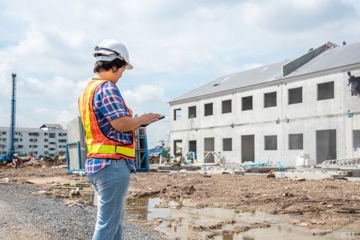 Asian woman civil construction engineer worker or architect with helmet and safety vest working and holding a touchless tablet computer for see blueprints or plan at a building or construction site