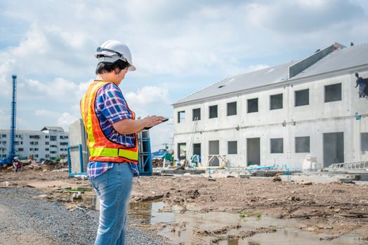 Asian woman civil construction engineer worker or architect with helmet and safety vest working and holding a touchless tablet computer for see blueprints or plan at a building or construction site