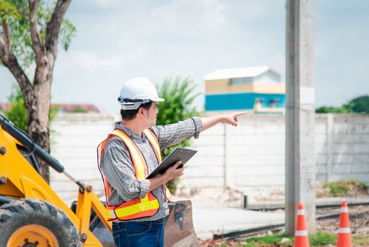 Asian man civil construction engineer worker or architect with helmet and safety vest working and holding a touchless tablet computer for see blueprints or plan at a building or construction site