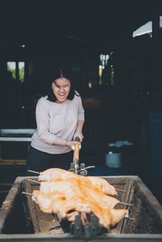 Asian woman chef cooking Barbecued Suckling Pig by roasting pork on charcoal for sale at Thai street food market or restaurant in Thailand