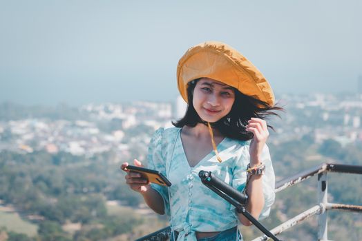 Asian pretty cute woman with hat relax and use smartphone at seaside city landscape viewpoint on mountain with happy and freedom emotion in concept travel, vacation, leisure in life