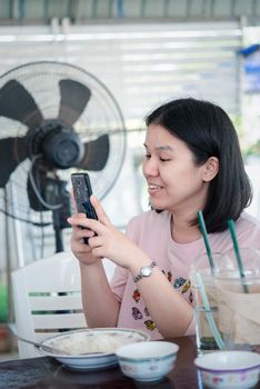 Asian woman foodstagramming use smartphone photographing a food on table in restaurant before eat for post and share on internet social media with fun and happy