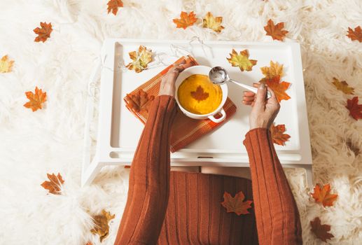 A female sits on a cream rug with a  bowl of spiced pumpkin rich and thick home made soup.  Seasonal fall maple leaves