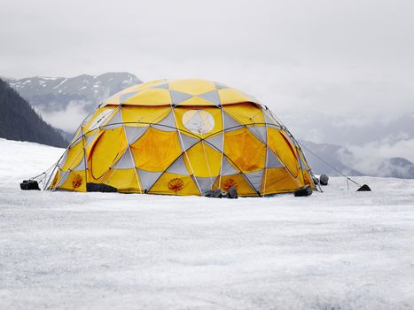 Orange-Grey Camping Tent On Icy Landscape. Rocky Mountains Ridges And Low Clouds At The Background