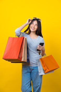 Attractive Asian smiling young woman Carrying a shopping coloful bag, mobile phone and sunglasses on aisolated yellow color background, copy space and studio, black friday season sale concept.