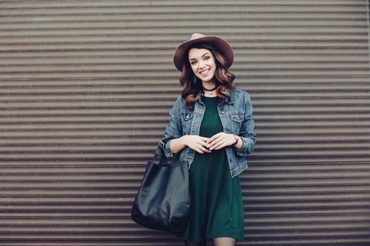 Stylish beautiful brunette woman in sunglasses, green dress, brown hat and big black bag, posing standing at street. Fashionable smiling girl leaning on wall. Concept of fashion and street trend look.